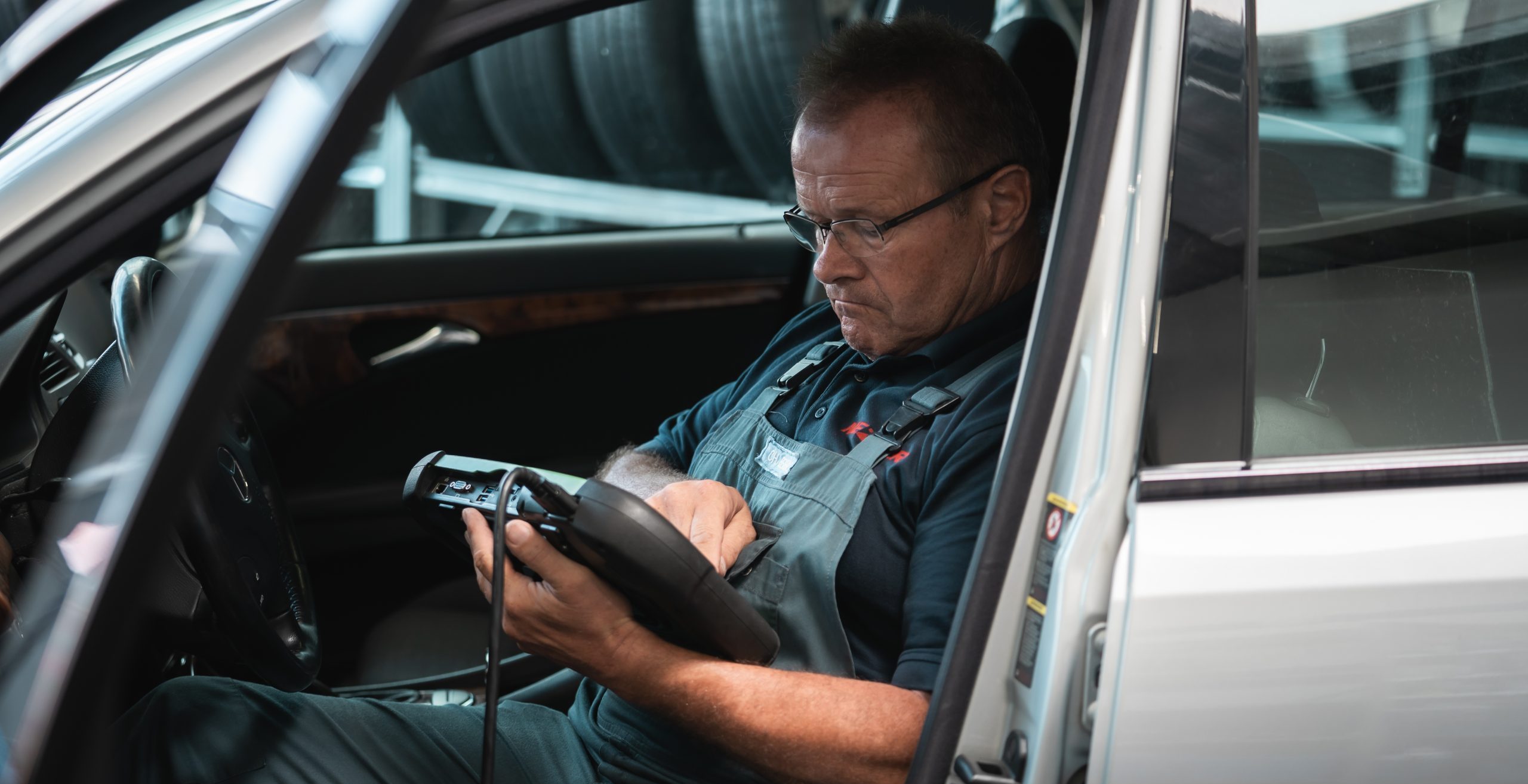 Ein KFZ-Mechatroniker sitzt auf dem Fahrersitz und hat ein Diagnosegerät in der Hand.